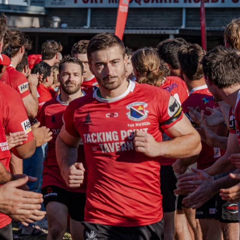 A player in a red Port Macquarie Pirates Rugby Union Club jersey running through a tunnel of teammates, featuring C2C Sport custom club apparel.